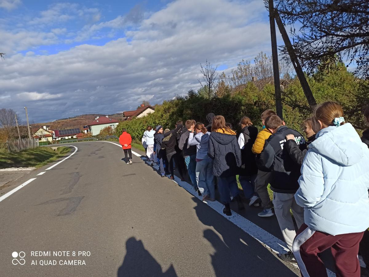 Grupa os&oacute;b idzie poboczem asfaltowej drogi na tle jesiennego krajobrazu z zabudowaniami i zachmurzonym niebem.